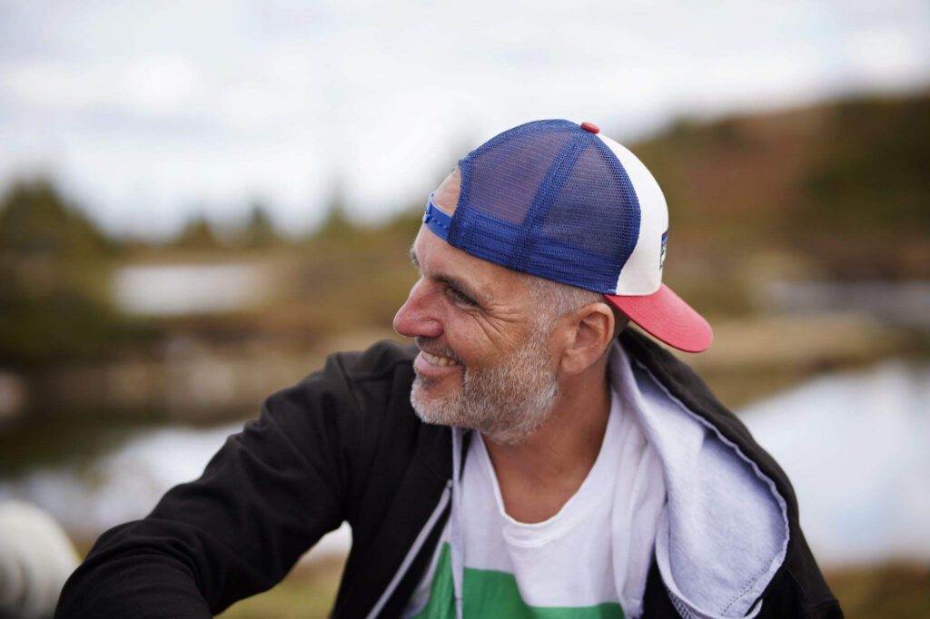 En medelålders man i profil med en keps kollar snett ifrån bilden med ett stort leenden. I bilden ser vi att fotografen heter Mattias Arbin. I bakgrunden skyms ett landskap.
