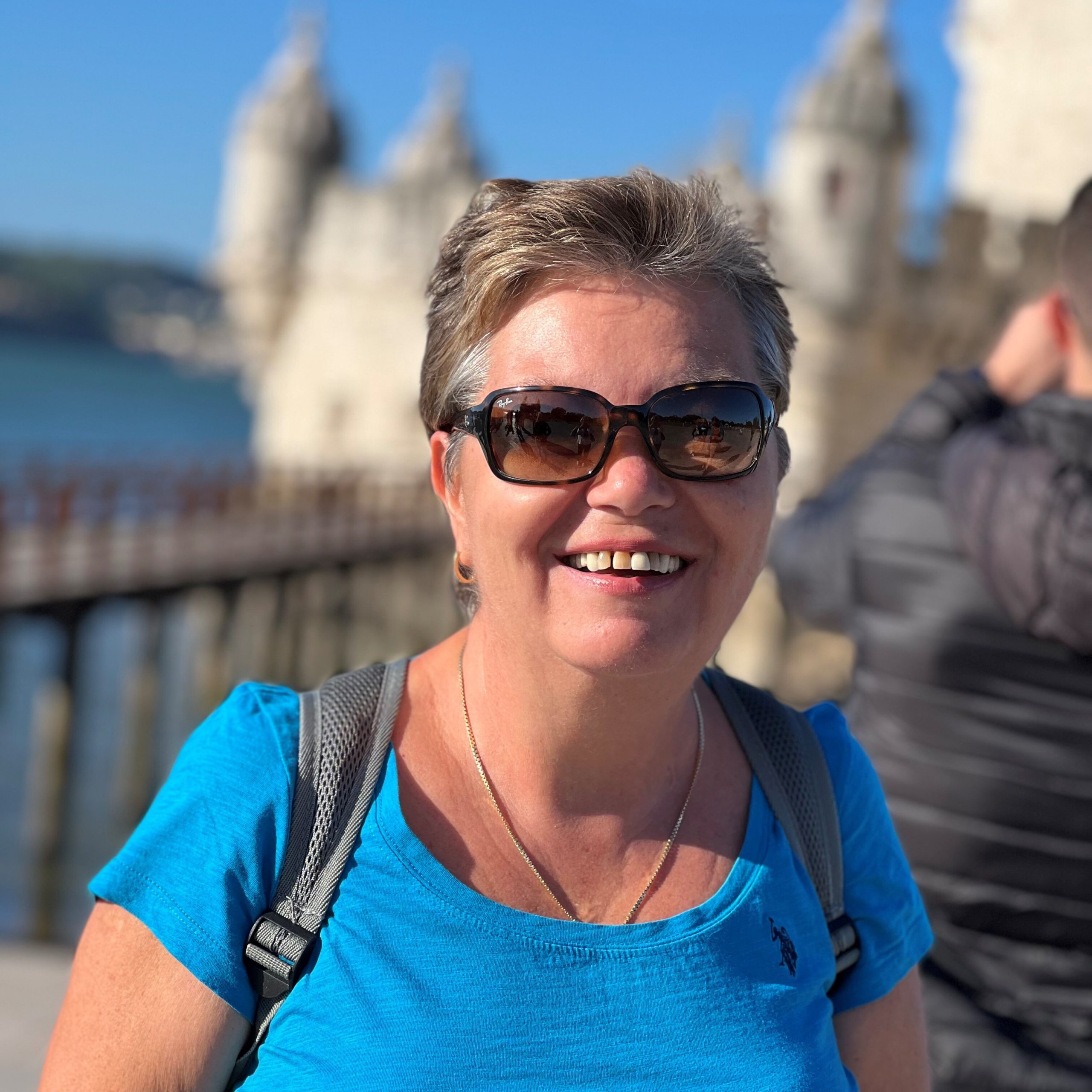 Ett personporträtt på en medelålders kvinna med solglasögon som ler och tittar in i kameran. I bakgrunden skymtar vi en männsika.