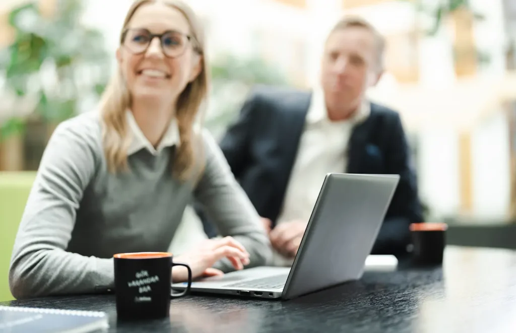 En kvinna med glasögon sitter i förgrunden och blickar bortåt samtidigt som hon lyssnar och antecknar på sin bärbara dator. I bakgrunden sitter även en man i kavaj och lyssnar.