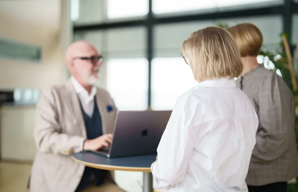 En äldre man sitter och skriver på sin dator samtidigt som han ler mot sina två kvinnliga medarbetare.