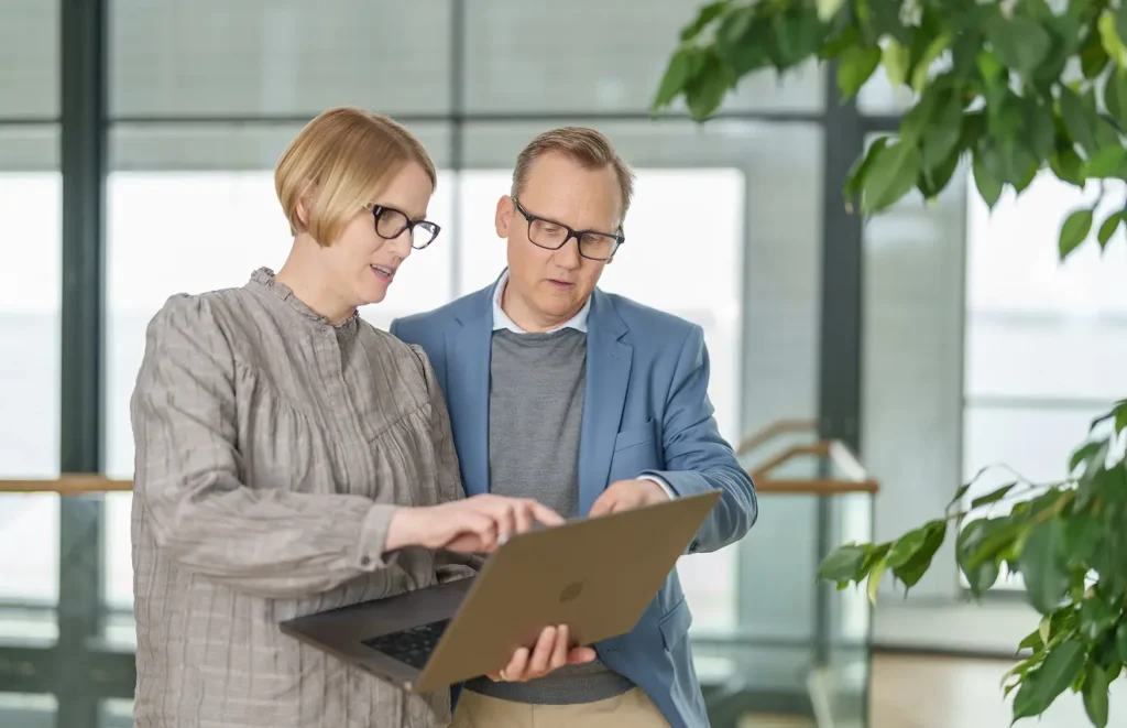 Två medelålders medarbetare, en man och en kvinna, står och diskuterar i en lobby. Kvinnan håller upp sin bärbara dator och pekar på sin skärm illustrativt för mannen.