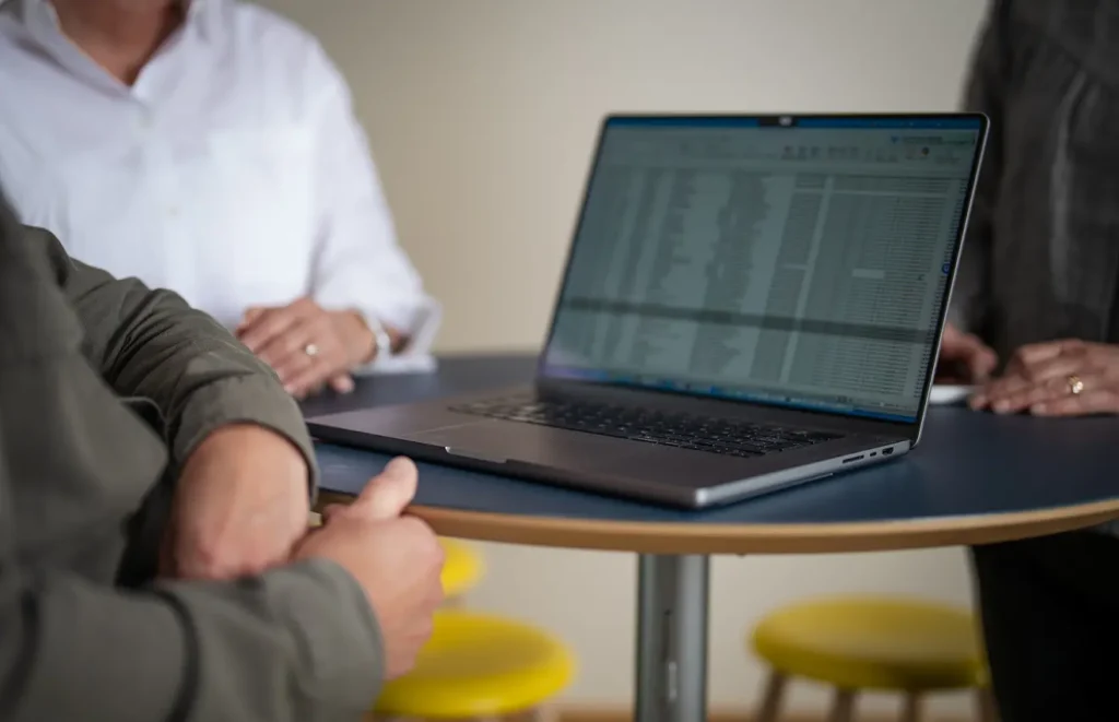 En bärbar dator står på ett bord som visar upp ett tekniskt dokument. Runt datorn sitter tre anonyma människor.
