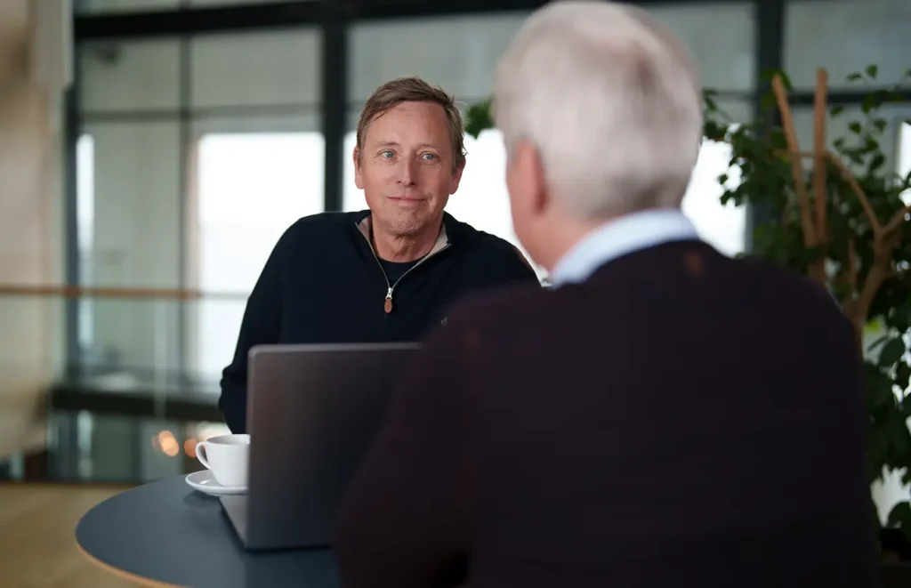 Två medelålders män sitter vid ett skrivbord och diskuterar med en dator emellan sig.