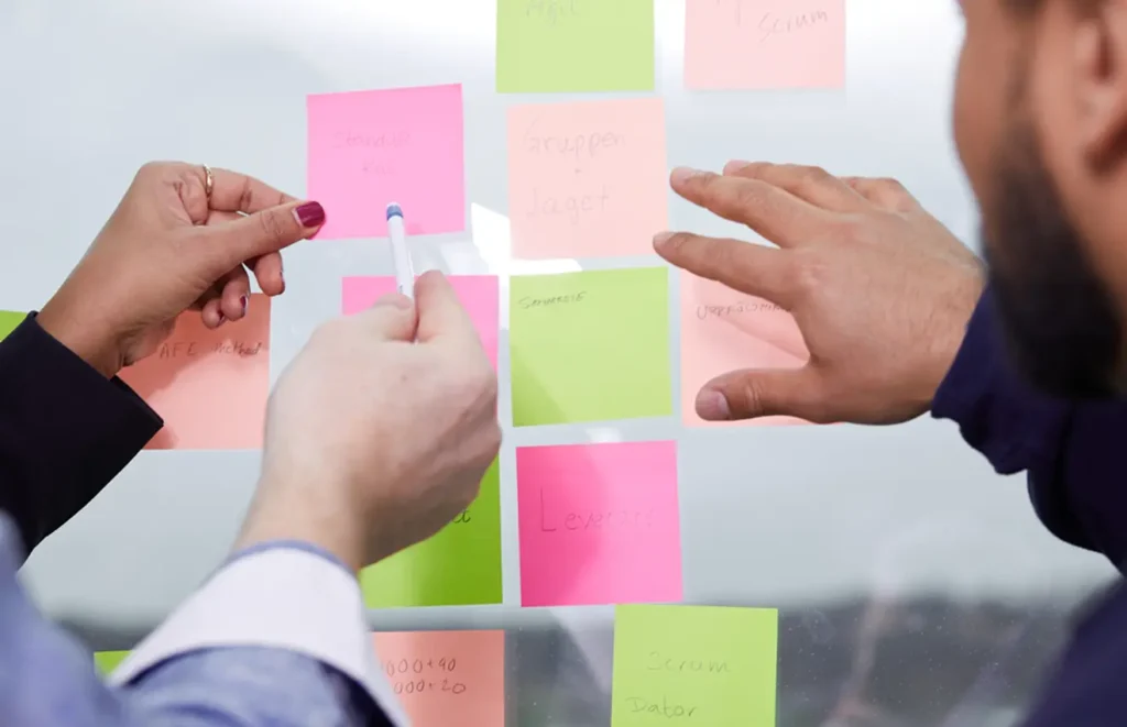 Ett flertal post-it-lappar sitter uppsatta på en vägg samtidigt som tre olika människors händer håller på och flyttar runt ordningen på lapparna