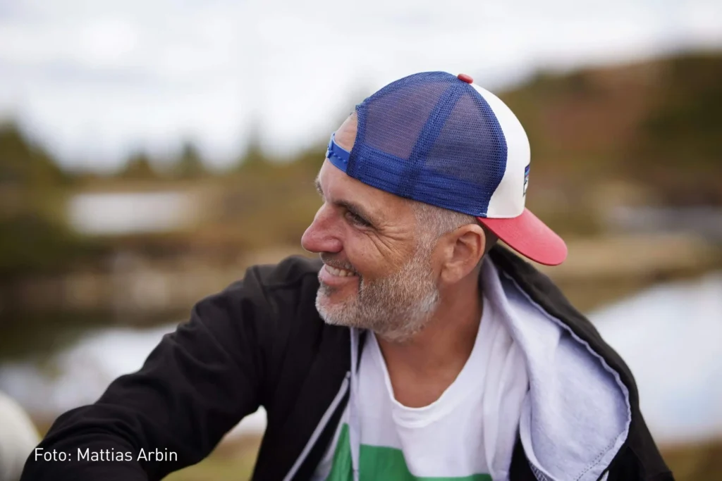 En medelålders man i profil med en keps kollar snett ifrån bilden med ett stort leenden. I bilden ser vi att fotografen heter Mattias Arbin. I bakgrunden skyms ett landskap.