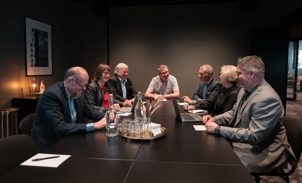 En grupp med medelålders kvinnor och män sitter samlade i ett konferensrum runt ett stort bord. Några har datorer framför sig, några har block och penna. De skrattar, ler och pratar med varandra. mitt på bordet står två vattenflaskor och bredvid står vattenglas.
