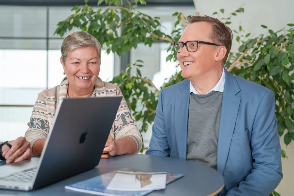 En medel ålderskvinna och en medelålders man ler och lyssnar på en man vars hand vi skymtar i bild. Vi ser även en dator som de är samlade runt.