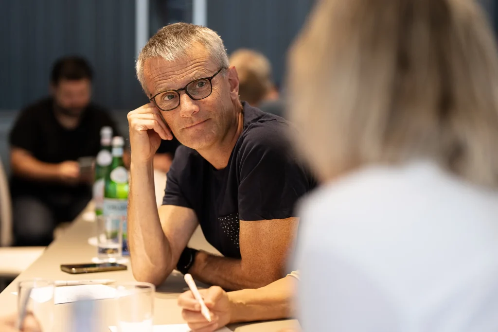 I förgrunden skymtar vi en kvinna med en vit tröja. Fokus i bilden är en medelålders man som lyssnar på kvinnan. Han har glasögon och en svart tröjan. I bilden skymtas även en arm och hand som noterar ner på papper. I bakgrunden kan vi också skymta två män som disskuterar.