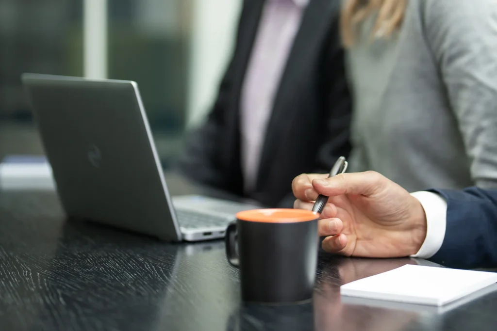 En närbild på en bärbar dator som står på ett skrivbord. I förgrunden syns en mans hand i kostym som håller en penna. I bakgrunden sitter ytterligare två anonyma människor, en man och en kvinna.