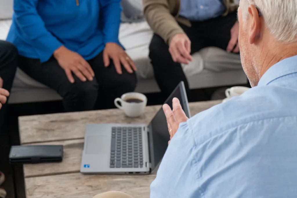 En grupp anonyma människor sitter runt ett soffbord. På bordet finns det en bärbar dator och kaffekopp. I förgrunden sitter det en medelålders man som gestikulerar med sina händer.