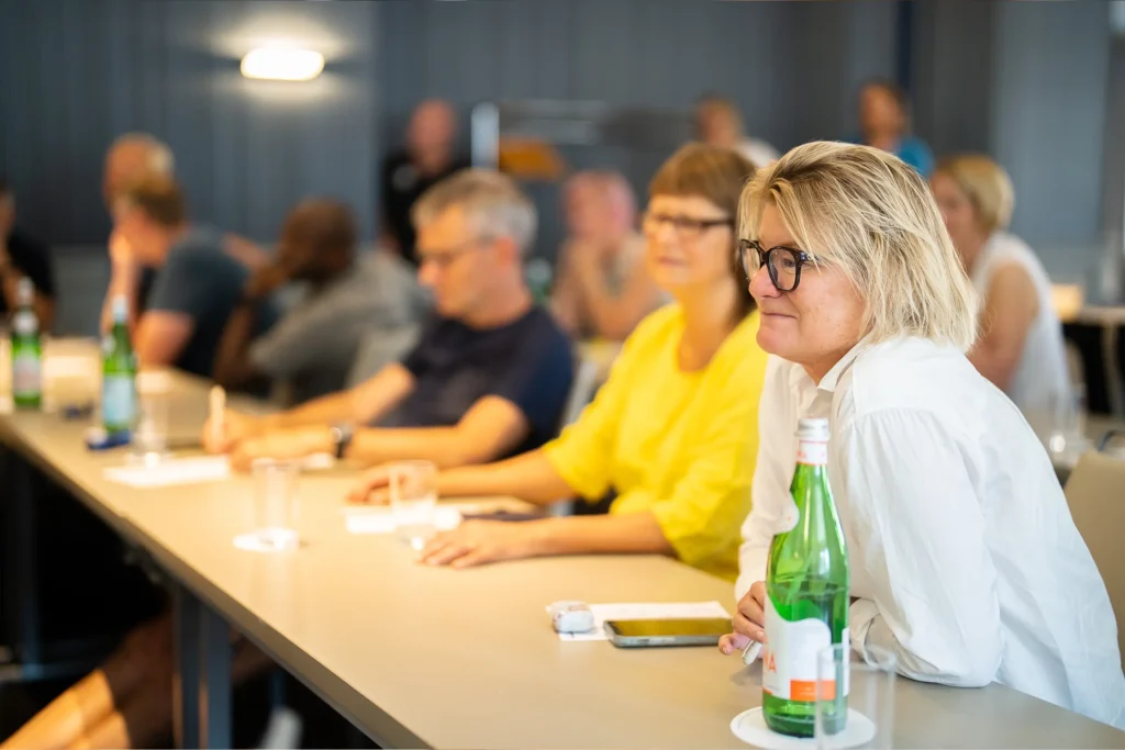 Vi ser en kvinna i förgrunden som lyssnar på en föreläsning. Framför sig har hon en vattenflaska, sin mobil och penna och papper. I bakgrunden skymtar vi flera andra människor som är ur fokus men som också lyssnar på föreläsningen. Alla sitter de vid bord med penna och papper framför dem.