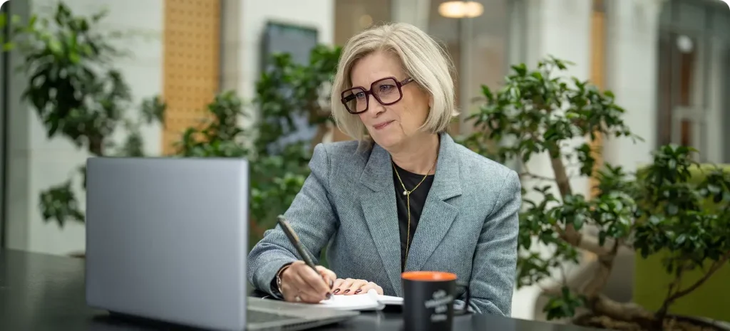 En medelålders kvinna som är proffesionellt klädd sitter med penna och block i handen kollar in i en datorskärm som står framför henne.