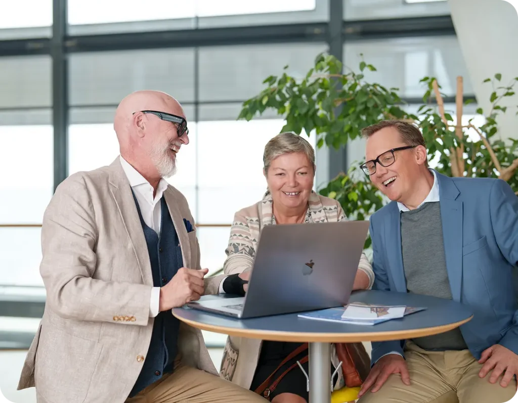 I mitten av bilden sitter en medelålders kvinna och två medelålders män sitter på var sin sida om henne. Alla tre tittar på en dator skärm som den ena mannen har. Alla är proffesionellt klädda. De ler stort och pratar med varandra.
