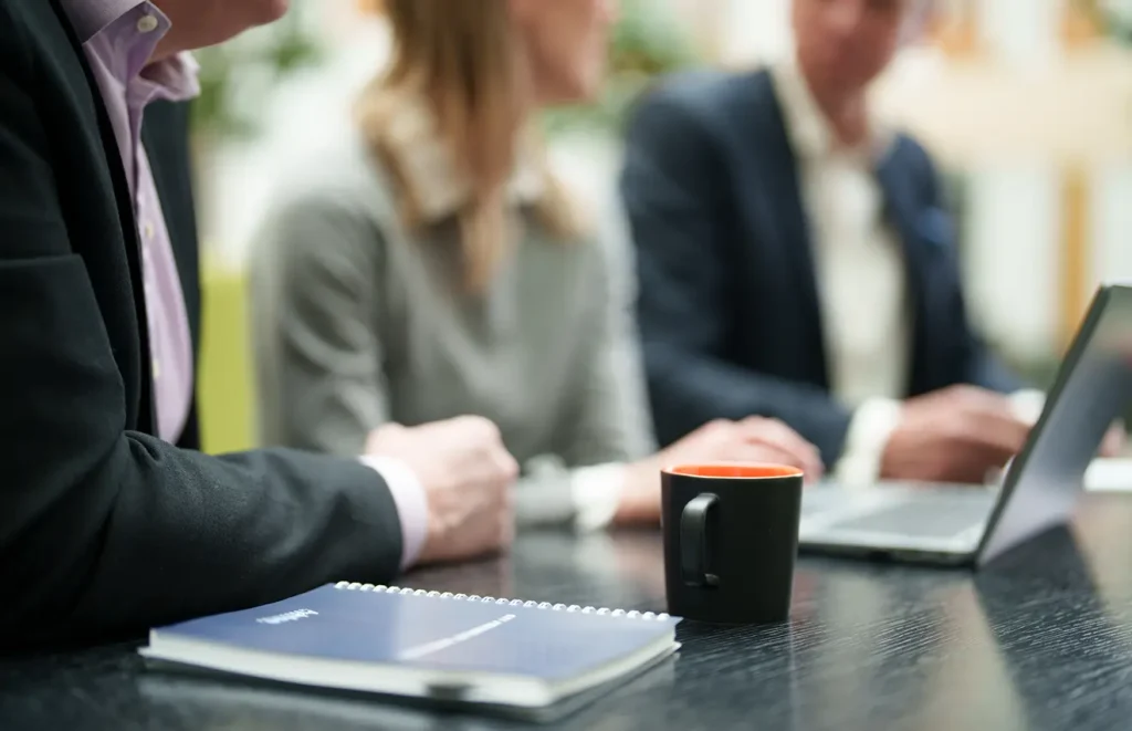 Tre anonyma, professionellt klädda, människor sitter vid ett skrivbord. På bordet finns det antecjningsblock, en kaffemugg och bärbar dator.