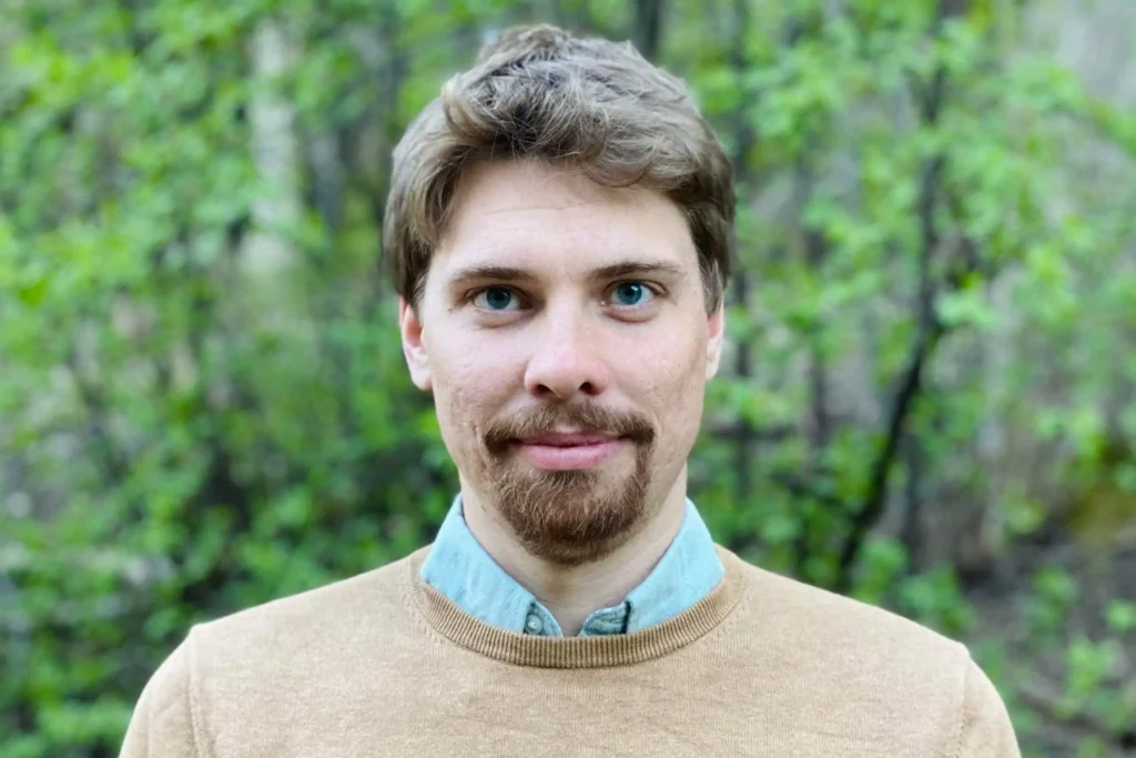 En personporträtt på en man med skägg och proffesionellt klädd som tittar in i kameran. Bakom skymtar vi grönska.