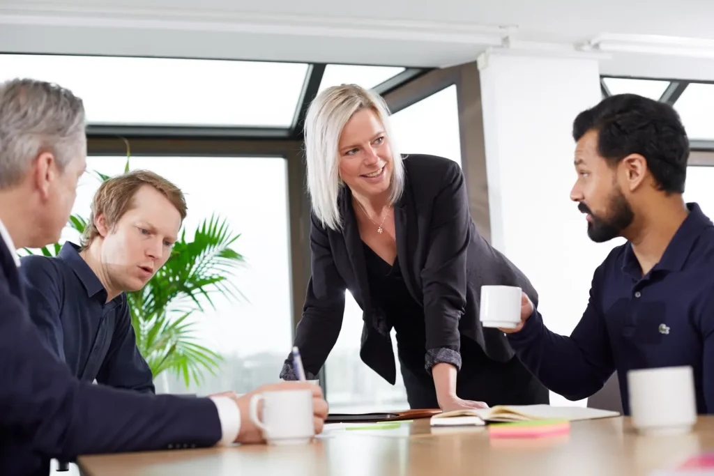 En kvinna i professionella kläder lutar sig över ett bord och pratar med en man till höger om henne. Till vänster sitter även två män som tittar på varandra och pratar.