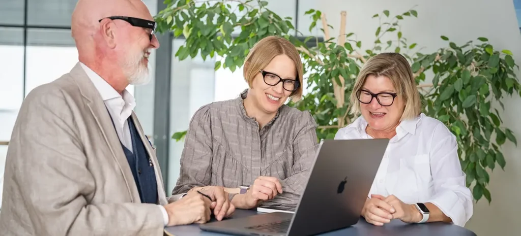 En medelålders man och och två medleålders kvinnor pratar, ler och tittar på en datorskärm.