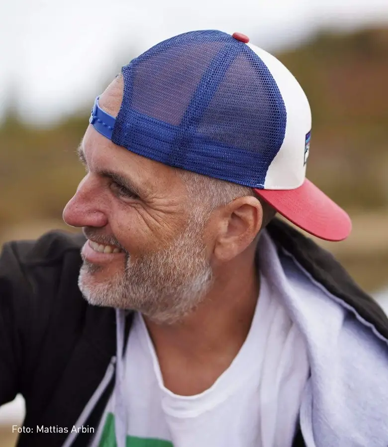 En medelålders man i profil med en keps kollar snett ifrån bilden med ett stort leenden. I bilden ser vi att fotografen heter Mattias Arbin.